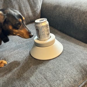 May include: A white can holder with a gray base and a silver can sitting on top. A brown and black dachshund is looking at the can holder.