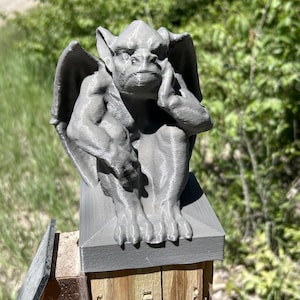 May include: A gray gargoyle statue with large wings, sitting on a gray square base. The statue has a detailed face, hands, and feet. The gargoyle is perched on a wooden post, with green foliage in the background.