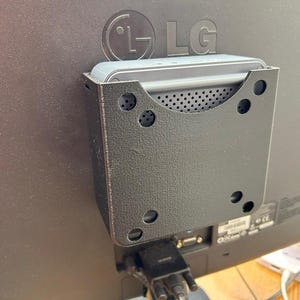 May include: A black plastic wall mount for a small electronic device, with a silver device attached to it. The mount has several holes for screws and is attached to the back of a black LG monitor.