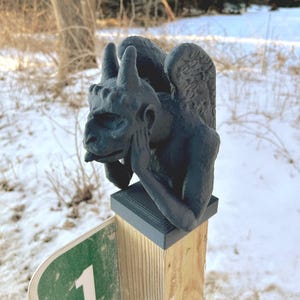 May include: A gray stone gargoyle sculpture with wings, sitting on a wooden post. The gargoyle is looking down with its head resting on its hand.