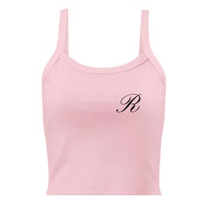 May include: A light pink tank top with thin straps. The top features a black cursive letter "R" on the front. The fabric appears to be a soft, ribbed material. The design is simple and minimalist.