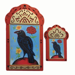 May include: Two red wooden frames with intricate cutouts, each featuring a black crow perched on a red orb. The larger frame has a blue background with yellow flowers and a red rose. The smaller frame has a red background with yellow flowers and a red rose.