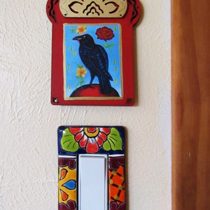 May include: A decorative light switch cover with a colorful floral design. The cover is made of ceramic and features a white light switch in the center. The switch cover is mounted on a white wall next to a wooden door.