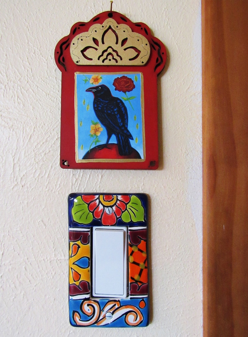 May include: A decorative light switch cover with a colorful floral design. The cover is made of ceramic and features a white light switch in the center. The switch cover is mounted on a white wall next to a wooden door.