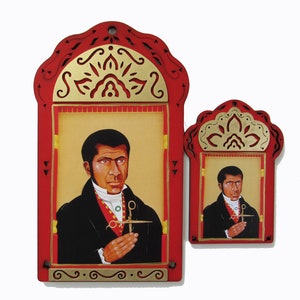 May include: Two red wooden framed images of a man with dark hair and a black suit holding a pair of scissors. The man is depicted in a painted portrait style. The frames have gold accents and intricate designs.