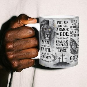 Op de afbeelding: Witte keramische mok met een leeuw en religieuze teksten in zwart en grijs. De mok bevat zinnen als "Man van Geloof" en "Pantser van God". De mok wordt vastgehouden door een persoon die een wit t-shirt draagt.
