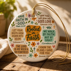以下が含まれることがあります： 中央に「Rooted in Faith」という文字が書かれ、その周囲に様々な聖書の一節とカラフルな花のイラストが描かれた円形の白いセラミック製オーナメント。上部の穴には金色の吊り下げ用紐が通されています。