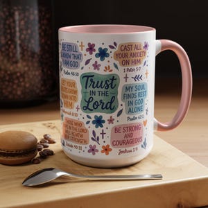 May include: White ceramic mug with a pink interior and handle, decorated with floral designs and inspirational text, including "Trust in the Lord." A spoon and a pastry are on a wooden surface.