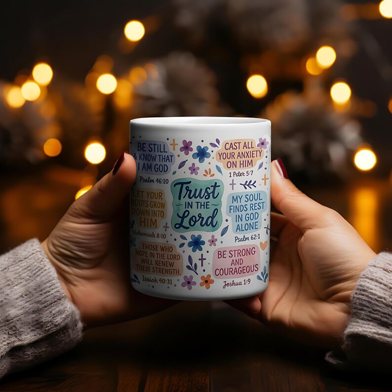 May include: White ceramic mug featuring floral and cross designs, and inspirational text including "Trust in the Lord" and Bible verses. The mug is held by hands, with a blurred background of warm lights.