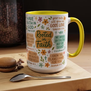 May include: White ceramic mug with a yellow interior and handle. The mug is decorated with floral designs and inspirational text, including "Rooted in Faith" and Bible verses. A spoon and a pastry are on a wooden surface.