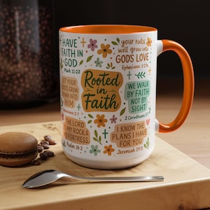 May include: White ceramic mug with an orange interior and handle. The mug is decorated with floral designs and inspirational text, including "Rooted in Faith" and Bible verses. A spoon and a pastry are on a wooden surface.