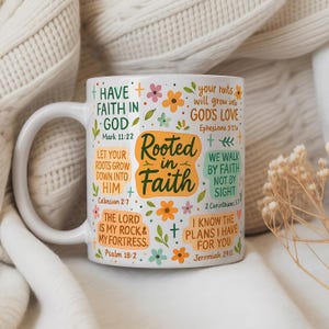 May include: White ceramic mug with a curved handle, featuring the text "Rooted in Faith" in an orange and green design. The mug is decorated with floral and cross motifs, and Bible verses. The mug is set against a soft, textured background.