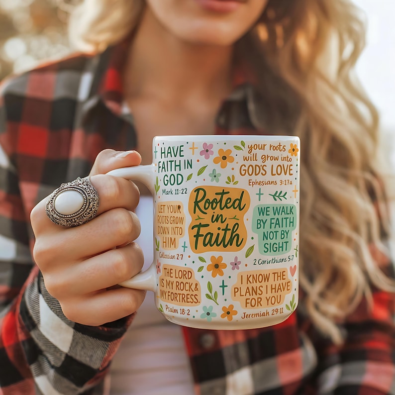 May include: White ceramic mug featuring the text "Rooted in Faith" and Bible verses, decorated with flowers. The mug has a curved handle and is held by a person wearing a ring.