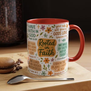 May include: A white ceramic mug with a red interior and handle. The mug is decorated with floral designs and inspirational text, including "Rooted in Faith" and Bible verses. The mug is on a wooden surface.