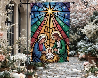 May include: A stained-glass style garden flag depicts the Nativity scene. Mary and Joseph flank the baby Jesus in a manger, beneath a radiant starburst. The flag features vibrant colors including blue, green, red, and yellow, and is displayed outdoors.