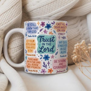 May include: White ceramic mug with a curved handle, decorated with floral accents and inspirational text. The mug features the words "Trust in the Lord" and various Bible verses in a colorful design. The mug is set against a soft, textured background.