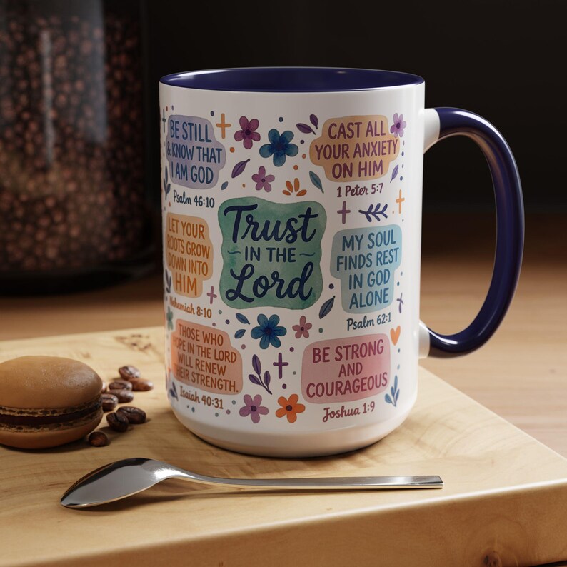 May include: A white ceramic mug with a navy blue handle and rim. The mug is decorated with colourful floral designs and inspirational text, including "Trust in the Lord" and Bible verses. A spoon and a macaron are on a wooden surface.