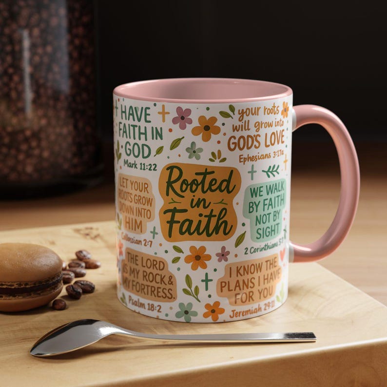 May include: A white ceramic mug with a pink interior and handle, decorated with floral and cross motifs. The mug features the text "Rooted in Faith" and various Bible verses. A spoon and a pastry are on a wooden surface.