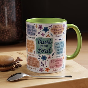 May include: A white ceramic mug with a green interior and handle, adorned with floral patterns and religious quotes. The mug displays the text "Trust in the Lord" and other inspirational phrases.