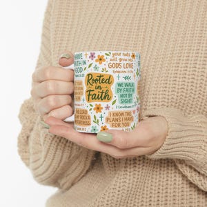 May include: White ceramic mug featuring the phrase "Rooted in Faith" and Bible verses. The mug is adorned with floral and cross motifs. The mug is held by a person.