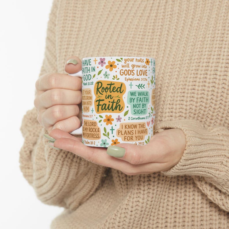 May include: White ceramic mug featuring the phrase "Rooted in Faith" and Bible verses. The mug is adorned with floral and cross motifs. The mug is held by a person.