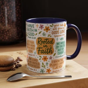 May include: A white ceramic mug with a navy blue interior and handle. The mug is decorated with floral and cross designs, and inspirational text including "Rooted in Faith". A spoon and a pastry are in the foreground.