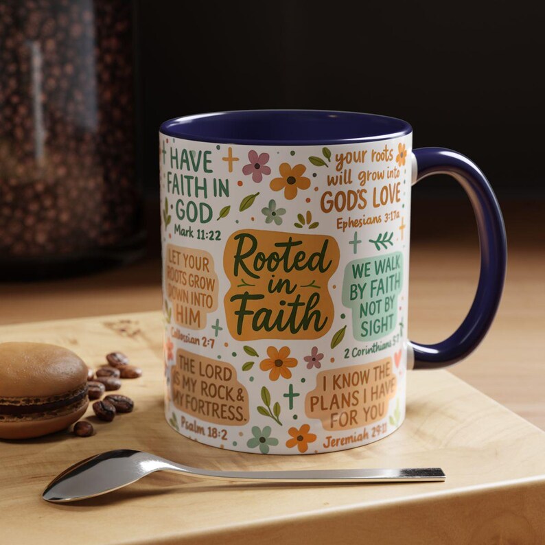 May include: A white ceramic mug with a navy blue interior and handle. The mug is decorated with floral and cross designs, and inspirational text including "Rooted in Faith". A spoon and a pastry are in the foreground.