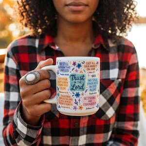 May include: White ceramic mug with a large handle, decorated with colourful floral designs and inspirational text. The mug includes phrases like "Trust in the Lord" and Bible verses. The person holding the mug is wearing a red and black plaid shirt.
