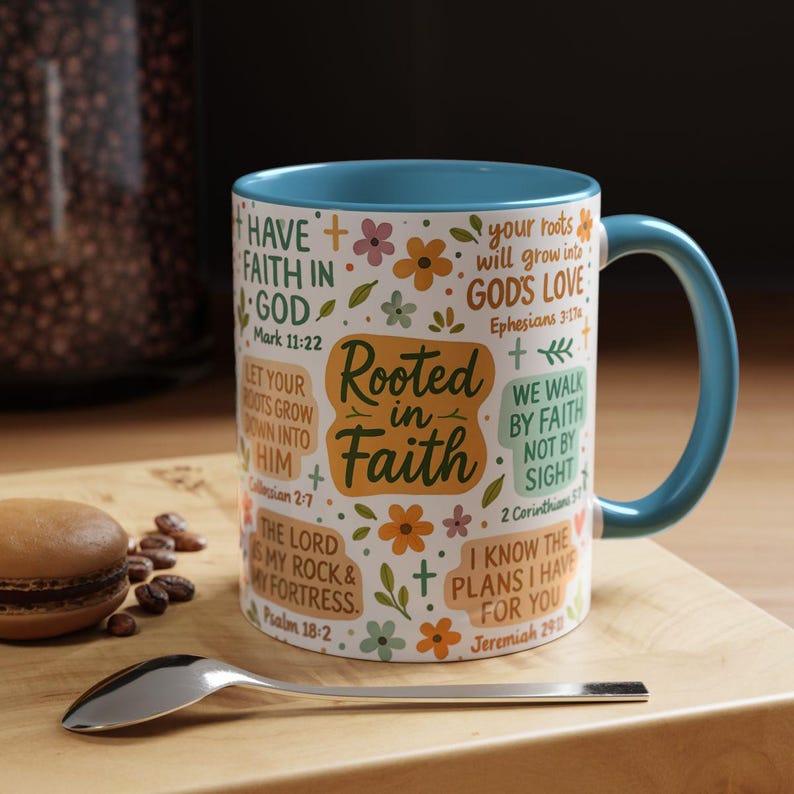 May include: A white ceramic mug with a teal interior and handle, featuring the text "Rooted in Faith" and various Bible verses. The mug is decorated with floral and cross motifs. A spoon and a pastry are in the foreground.