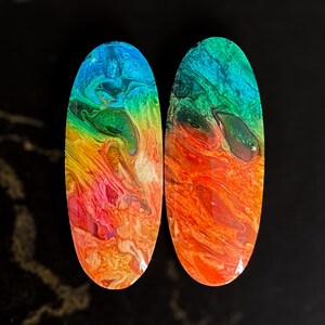 May include: Two oval-shaped, colorful resin earrings. The earrings feature a vibrant blend of colors, including blue, green, yellow, orange, and pink, creating a marbled effect. The earrings are set against a dark background.
