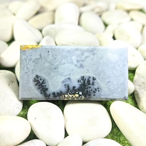 Op de afbeelding: Een rechthoekige, gepolijste edelsteen met een lichtgrijs en wit patroon, met zwarte dendritische insluitsels. De steen heeft een gele en gouden accent op één rand en wordt getoond op een bed van witte stenen.