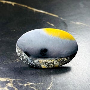 May include: An oval-shaped stone with a smooth, polished surface. The stone features a gradient of colors, including gray, yellow, and black. The bottom edge has a textured, speckled pattern. This unique stone could be used for jewelry or decorative purposes.