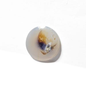 May include: A round, white agate cabochon with brown and black dendritic inclusions. The stone has a smooth, polished surface.