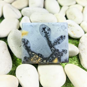 May include: Square gemstone with a pale blue background and black dendritic patterns. The stone has yellow and gold accents. The gemstone is surrounded by white stones on a green surface.