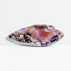Könnte beinhalten: Ein polierter, tropfenförmiger Edelstein mit einer Mischung aus Farben. Der Stein hat eine Basis aus lila und rosa Farbtönen mit Bereichen in Orange und Braun. Weiße, federartige Muster sind an den Rändern sichtbar.