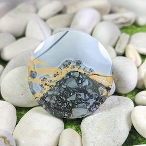 Pode incluir: Uma pedra preciosa redonda e polida com um padrão único. A pedra apresenta uma mistura de cores cinza, preto e bege, criando um design semelhante a uma paisagem. A pedra é rodeada por seixos brancos, sugerindo um ambiente natural.