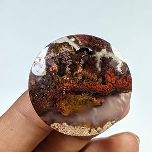 Puede incluir: Una piedra de ágata redonda y pulida con un patrón único de colores marrón, rojo, blanco y amarillo. La piedra tiene una superficie lisa y pulida y una forma natural y orgánica.