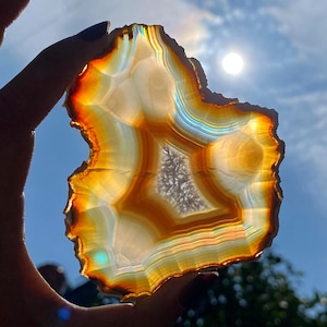 May include: A backlit agate slice, displaying layers of orange, yellow, and white. The natural stone has a central crystalline area and is held up against a bright sky, emphasising its vibrant colours and unique patterns. The stone's measurements are in centimetres.