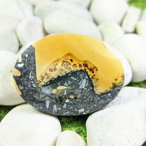 May include: Oval-shaped polished stone with a mix of colors. The top portion is a warm yellow, transitioning to a dark gray and speckled base. The stone is surrounded by white pebbles, creating a natural aesthetic.