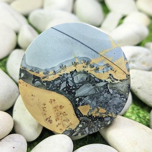 May include: A circular polished stone with a landscape design. The stone displays a mix of colours, including light blue, tan, black, and grey, reminiscent of a natural landscape. The stone is surrounded by white pebbles.