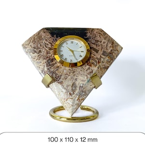 Puede incluir: Un reloj de cuarzo decorativo con una base de piedra en forma de diamante. El reloj tiene un bisel y manecillas doradas, con números romanos en la esfera blanca. La base es una mezcla de tonos marrón, beige y negro, sostenida por un soporte de anillo dorado. Dimensiones: 100 x 110 x 12 mm.
