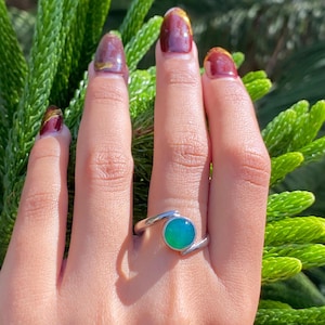 Peut inclure: Une bague en argent avec une pierre précieuse verte sertie dans une bande incurvée. La bague est sur le doigt d'une personne.