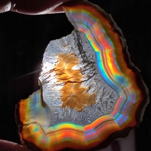 May include: A translucent, irregularly shaped stone with a rainbow-colored edge. The interior has a light brown, textured pattern. The stone is held against a dark background, highlighting the vibrant colors.