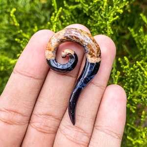 May include: A black and brown spiral-shaped ear gauge made of wood. The gauge has a smooth, polished finish.