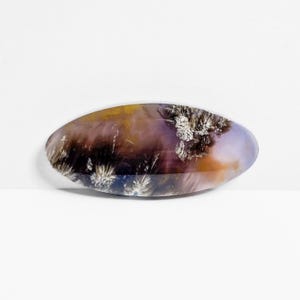 May include: An oval-shaped gemstone with a polished surface. The stone features a blend of colors, including purple, brown, yellow, and white, with a unique, natural pattern. The gemstone is set against a white background.