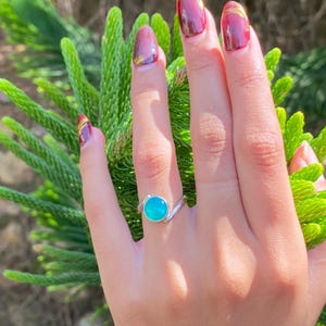 Puede incluir: Un anillo de plata con una piedra redonda de color turquesa brillante. El anillo está en una mano con uñas cuidadas. El fondo presenta follaje verde.