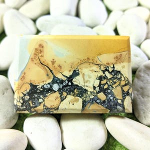 Puede incluir: Una gema rectangular con un patrón único de colores beige, negro y gris. La piedra tiene una superficie pulida y un aspecto natural y terroso. El diseño se asemeja a un paisaje con grietas y variaciones.