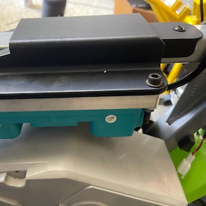 May include: A black metal component with a rectangular shape, secured with bolts, sits atop a teal-colored rectangular base. The assembly is mounted on a light gray surface, with a glimpse of a yellow structure in the background.