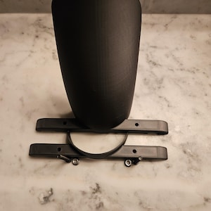 May include: A black, 3D-printed component, possibly for a vehicle or machine. It has a curved, cup-shaped section above a base with a semi-circular band and two parallel bars. The object is placed on a surface with a marble effect.