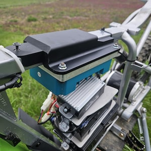 Puede incluir: Primer plano de un cuadro de bicicleta eléctrica con una caja de batería negra y una base azul. El cuadro es plateado y los componentes están montados sobre una plataforma de madera. El fondo muestra un campo y edificios.